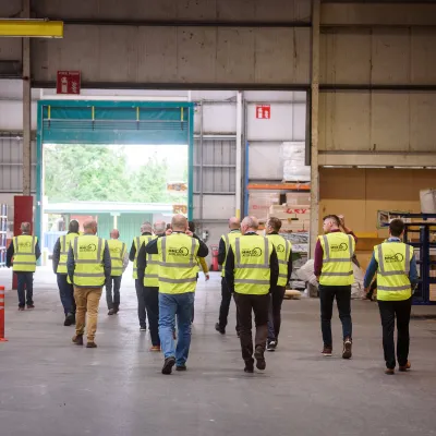 A group of people wearing high-visibility vests with the MMC Accelerate logo walking through a large warehouse towards an open entrance.