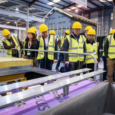 Participants in high-visibility vests and helmets observing a construction demonstration inside a manufacturing facility.