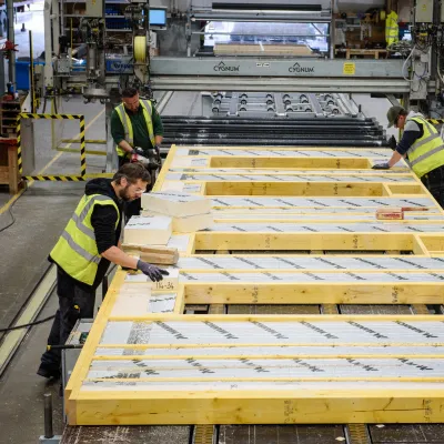 Workers in high-visibility vests assembling insulated timber wall panels inside a modern offsite construction factory.