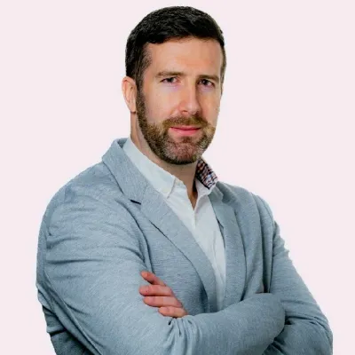 Professional portrait of person in light blue blazer with arms crossed against pale pink background.