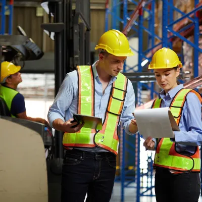 Warehouse workers in yellow hard hats discussing operations near blue storage racks.