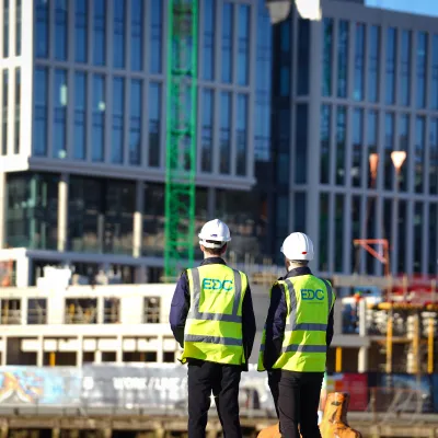 Two people wearing hard hats and high-visibility vests with the EDC logo, observing a modern glass building under construction.