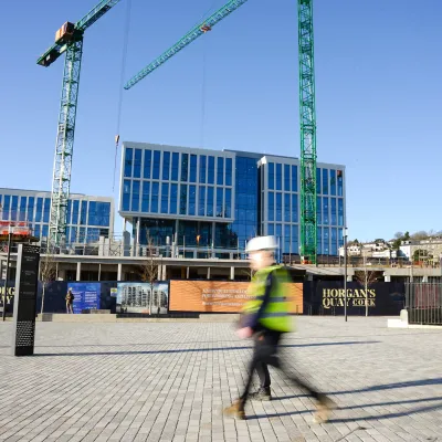 Construction site with cranes and modern glass buildings under development, with a person in safety gear walking in the foreground.