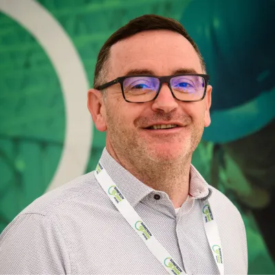 Person wearing glasses and a white shirt with a branded lanyard, standing in front of a green background.