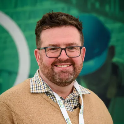 Person wearing glasses, a checked shirt, and a tan jumper with a branded lanyard, standing in front of a green background.