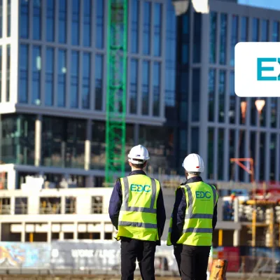 Two people wearing hard hats and high-visibility vests with the EDC logo, observing a modern glass building under construction.
