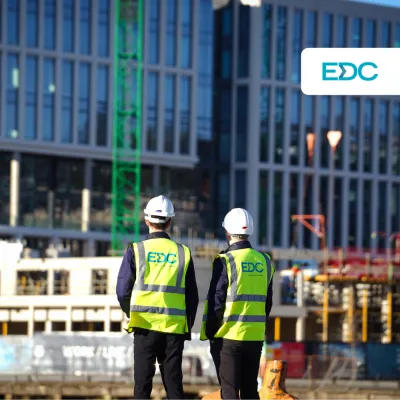Two people wearing hard hats and high-visibility vests with the EDC logo, observing a modern glass building under construction.