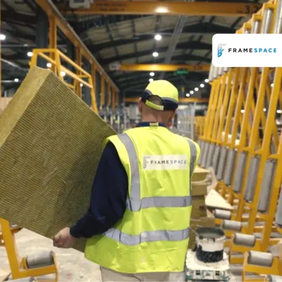 Worker in yellow hard hat and safety vest carrying insulation material in a warehouse.