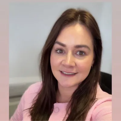 Woman with long brown hair smiling, wearing a pink top against a light background.