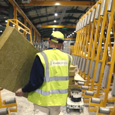 Person in safety vest working in warehouse with packages on yellow shelving racks.