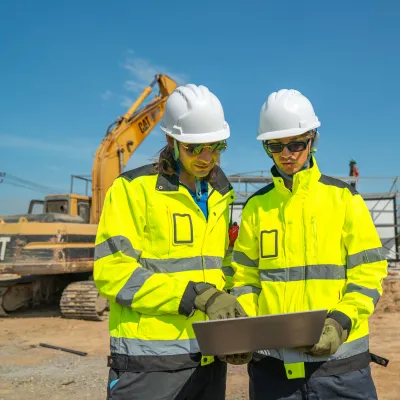 Two construction workers in hard hats and high-vis jackets reviewing plans at a building site.