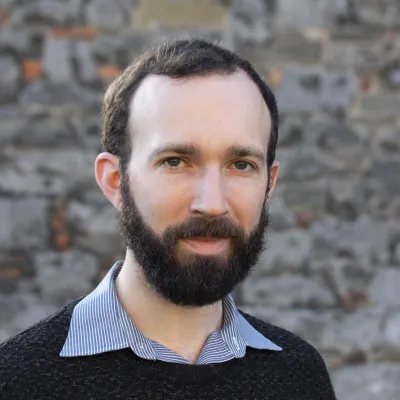 Person with a beard and short hair wearing a dark sweater and striped shirt, standing in front of a stone wall.