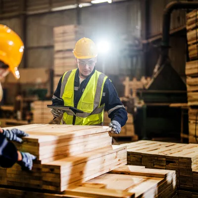 Worker in yellow hard hat and safety vest examining wooden planks in a warehouse setting.