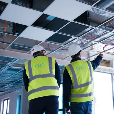 Two people wearing safety helmets and high-visibility vests inspecting ceiling installations inside a building under construction.