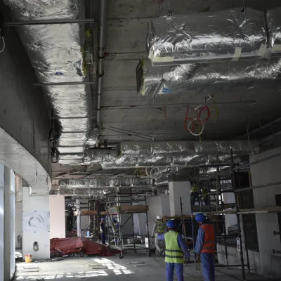 Construction site interior showing exposed ceiling with pipes and ductwork, and unfinished concrete walls.