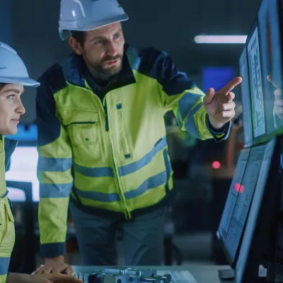 Two workers in hard hats and safety vests examining a digital screen in an industrial setting.