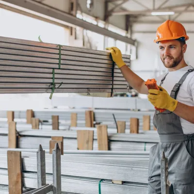 Construction worker in orange helmet reviewing documents in warehouse with stacked building materials.