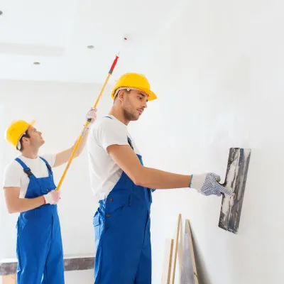 Two workers in yellow hard hats and blue overalls plastering a white interior wall.