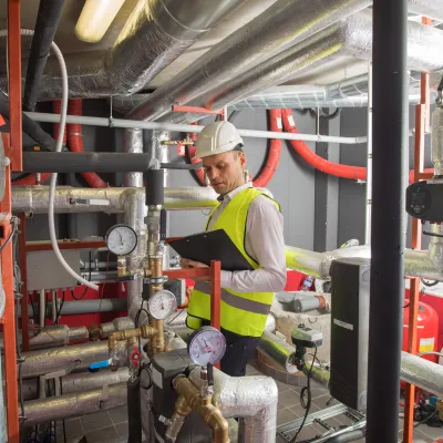Construction worker in safety vest and helmet working on pipes and valves in industrial setting.