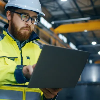 Construction worker in hard hat and safety vest working on laptop inside industrial facility.