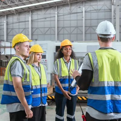 Three workers in hard hats, safety vests, and protective gear discussing at an industrial facility.