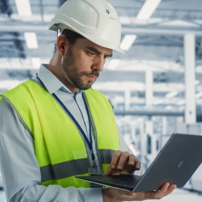 Worker in hard hat and safety vest using laptop in industrial facility.