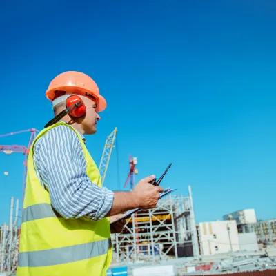 Construction worker in safety vest and helmet using tablet at building site against blue sky.