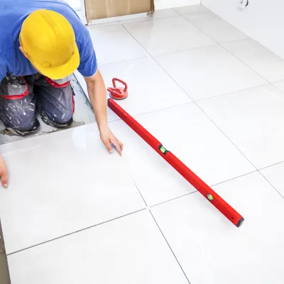 Construction worker in yellow hard hat laying white tile flooring with a red leveling tool.