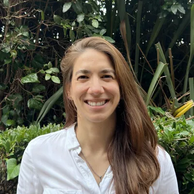 Woman with long brown hair in white shirt smiling in front of lush tropical plants.
