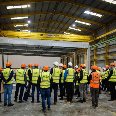 Group of workers wearing safety vests and hard hats gathered in an industrial warehouse facility.