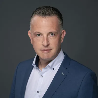 Professional portrait of person in navy blue suit with white shirt against dark background.