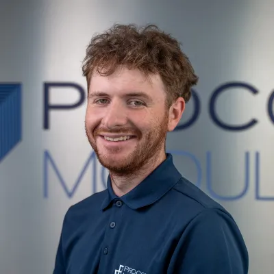 Smiling person with curly hair wearing a blue polo shirt in an office setting.