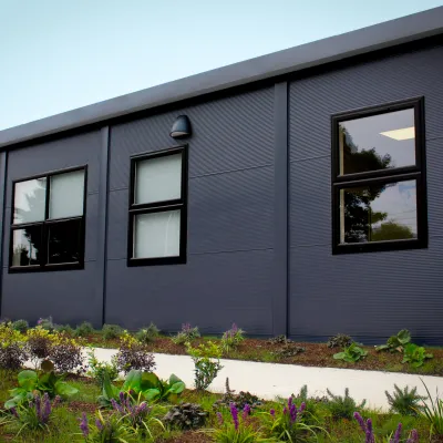 A modern single-story dark blue building with large windows surrounded by landscaped garden beds.