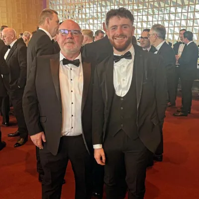 Two men in tuxedos posing on red carpet at formal event with audience in background.
