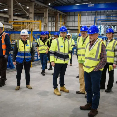 Group of people in hard hats and hi-vis vests touring a manufacturing facility with blue overhead crane.