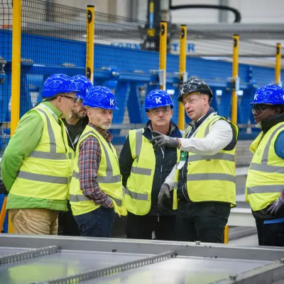 Workers in hi-vis vests examining materials in an industrial manufacturing facility with blue machinery.