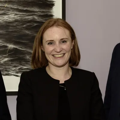 Person with reddish hair wearing a black outfit smiling, standing in front of artwork.