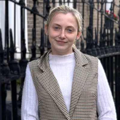 Person in a white turtleneck and checkered vest standing on a city street with black railings.