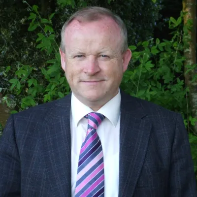Person in dark suit with purple striped tie standing outdoors against green foliage.