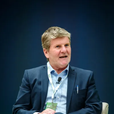 Person in a dark suit with a lanyard sitting on stage against a blue backdrop.