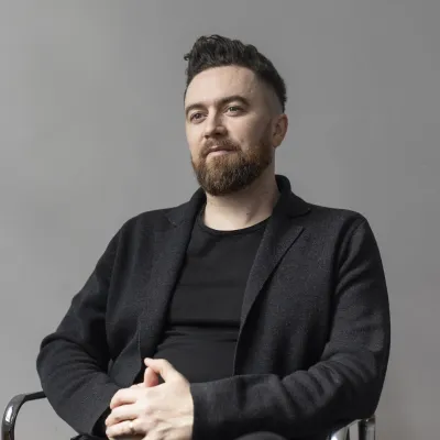 Person in black blazer and shirt seated against gray background.