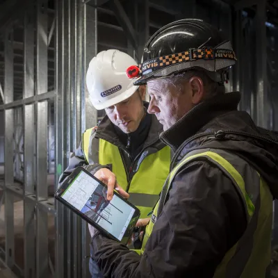 Two construction workers in safety gear examining a document inside an unfinished building.