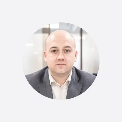 Professional headshot of person in dark suit with light shirt against neutral background.