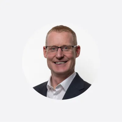 A smiling person with glasses wearing a dark suit and white shirt in a circular portrait format.