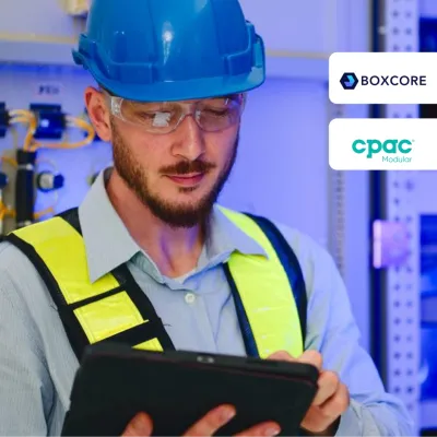Worker in blue hard hat and safety vest using tablet in industrial setting.