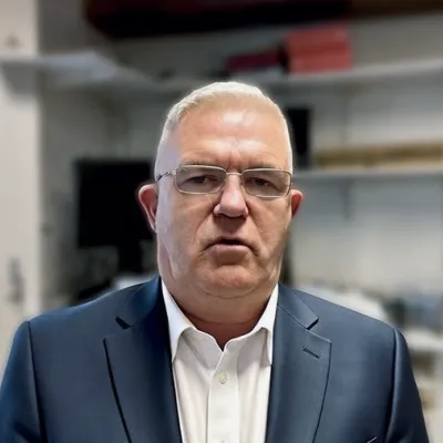 A person in a navy suit and white shirt wearing glasses against a blurred background.