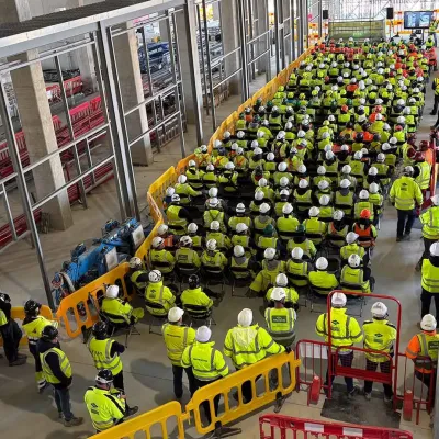 A large indoor gathering of workers in yellow safety vests and hard hats in a warehouse-like facility.