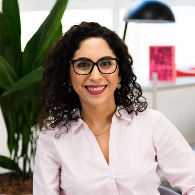 Woman with curly hair and glasses smiling in an office setting with plants visible.