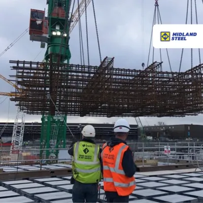 Construction workers in hard hats watching a large metal grid structure being lifted by crane at a building site.