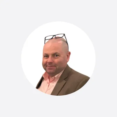 Man in brown suit jacket with glasses positioned on top of head, circular profile photo.